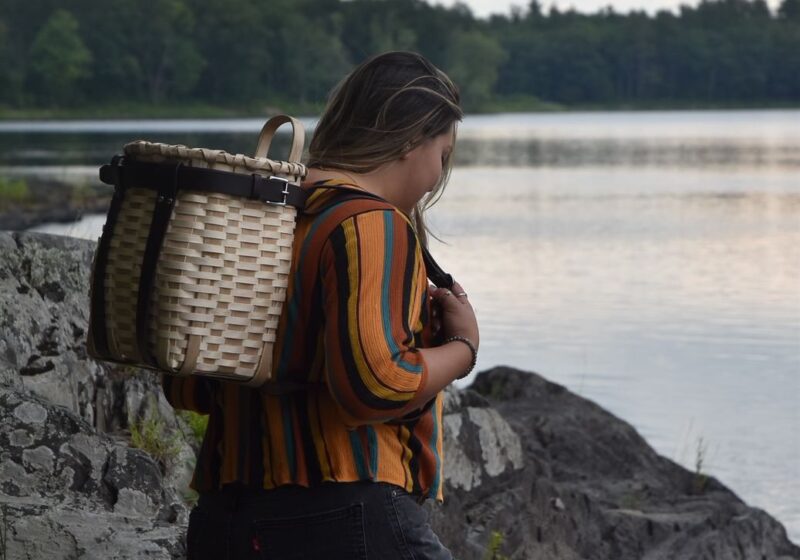 A woman wearing a pack basket stands on rocks at the edge of a lake