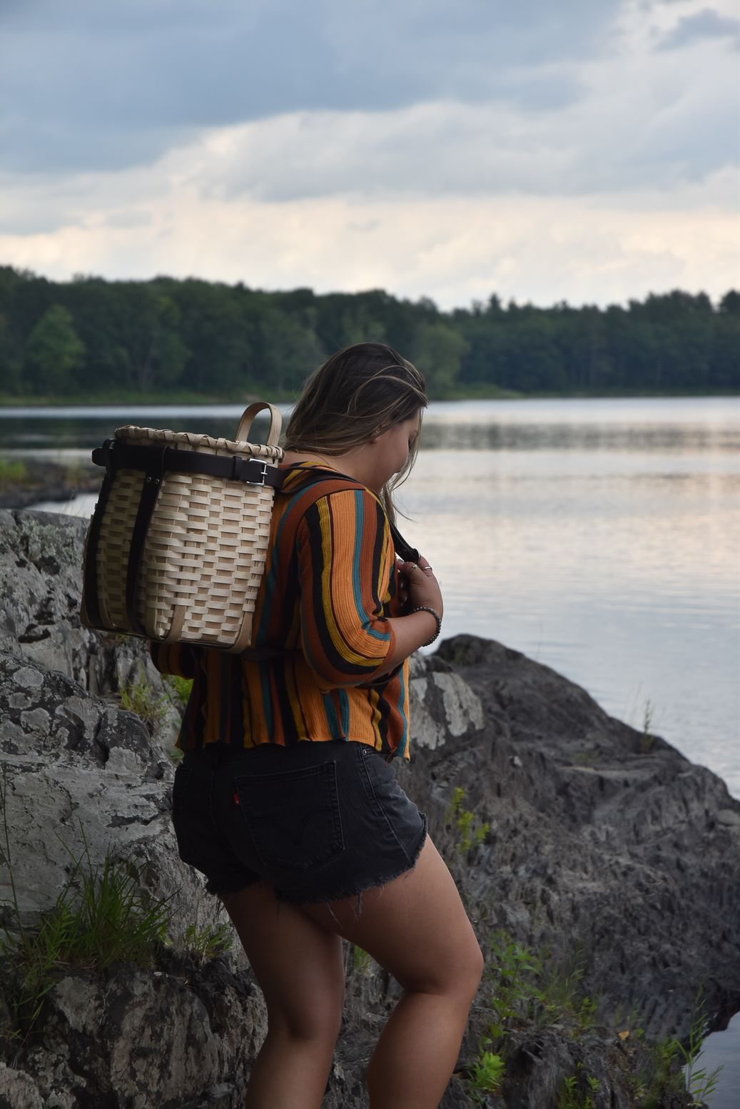 A woman wearing a pack basket stands on rocks at the edge of a lake