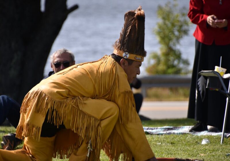 A dancer in fringed regalia and headdress kneels during a ceremony