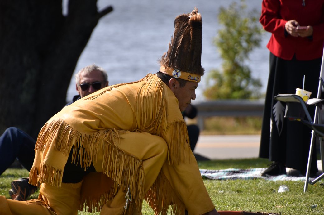 A dancer in fringed regalia and headdress kneels during a ceremony
